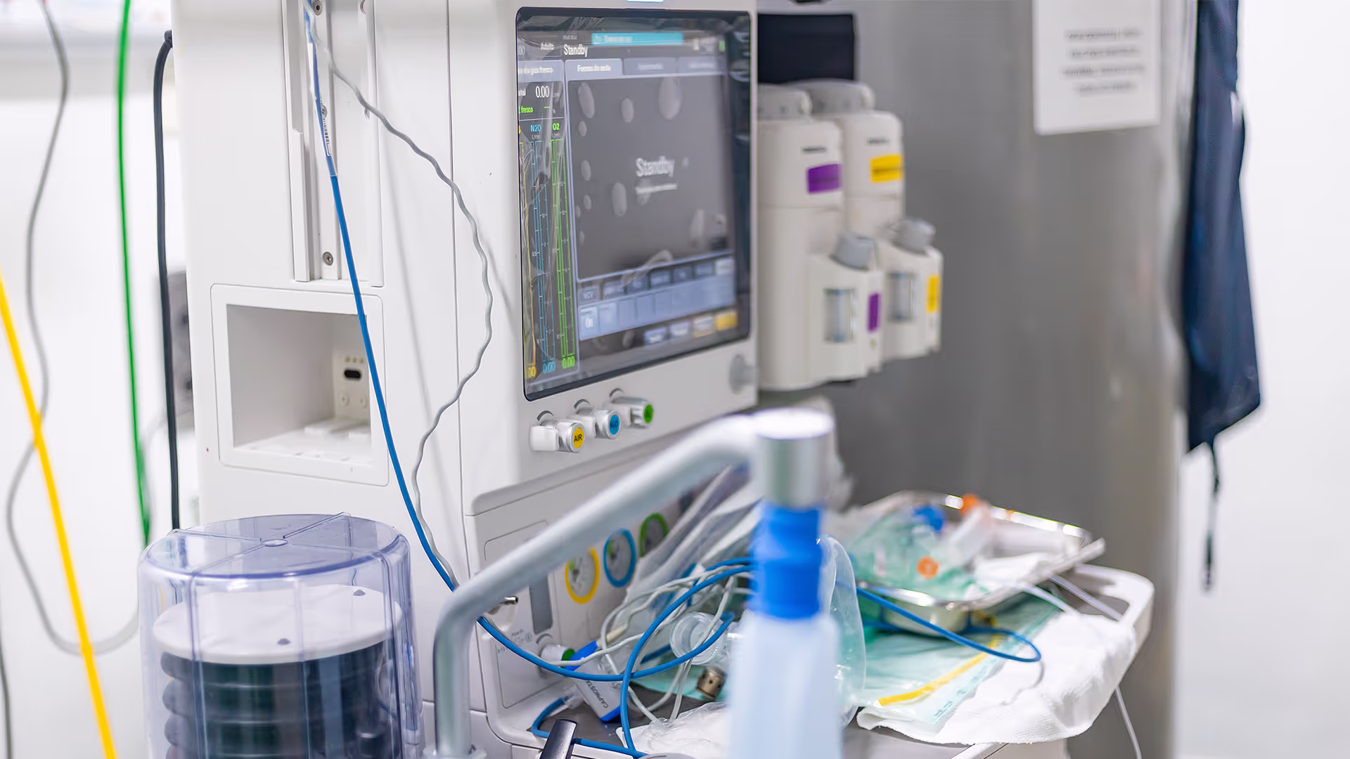 Equipamento médico em um ambiente cirúrgico, incluindo um monitor com gráficos e medições, frascos de anestesia acoplados e uma mesa auxiliar com materiais cirúrgicos e fios conectados. O cenário reflete a alta tecnologia utilizada em procedimentos médicos, garantindo precisão e segurança no tratamento dos pacientes.