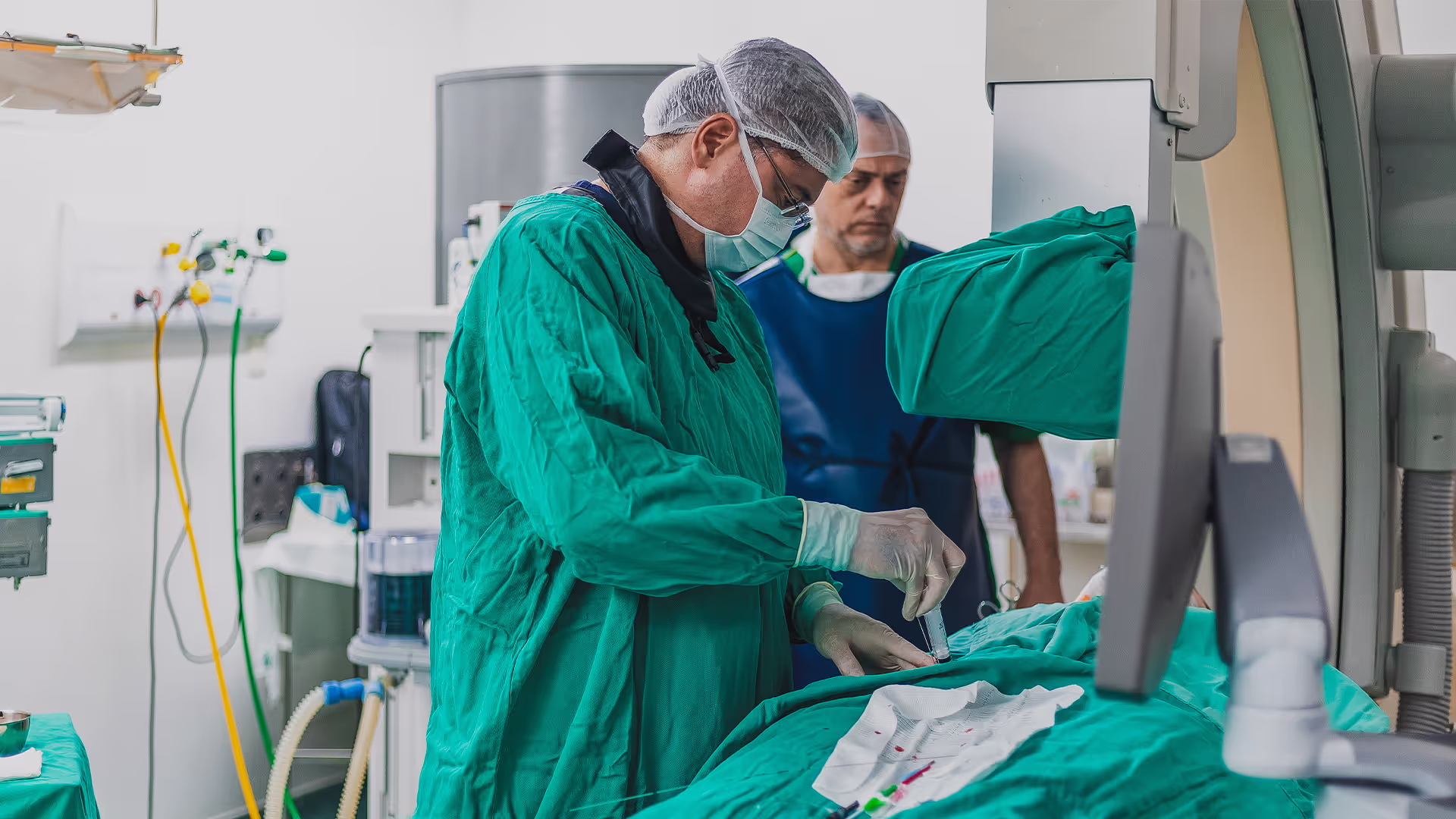 Médico em centro cirúrgico realizando um procedimento minimamente invasivo. Vestindo avental estéril, luvas, máscara e touca, ele manuseia um cateter sobre a mesa cirúrgica. Ao fundo, outro profissional de saúde, utilizando colete de proteção radiológica, acompanha o procedimento. O ambiente é equipado com monitores e aparelhos médicos de alta tecnologia, indicando um procedimento de eletrofisiologia ou intervenção cardiovascular.